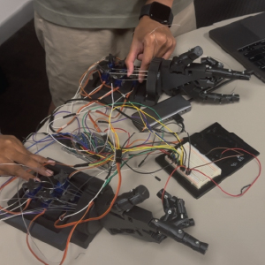 A person interacts with two robotic hand prototypes placed on a table, each connected to multiple servo motors and tangled wiring. The hands are mounted on black bases and linked to a breadboard and microcontroller setup. One hand appears to mirror or respond to the other, showing the gesture-mimicking system in development. A laptop sits nearby, used for programming or control.